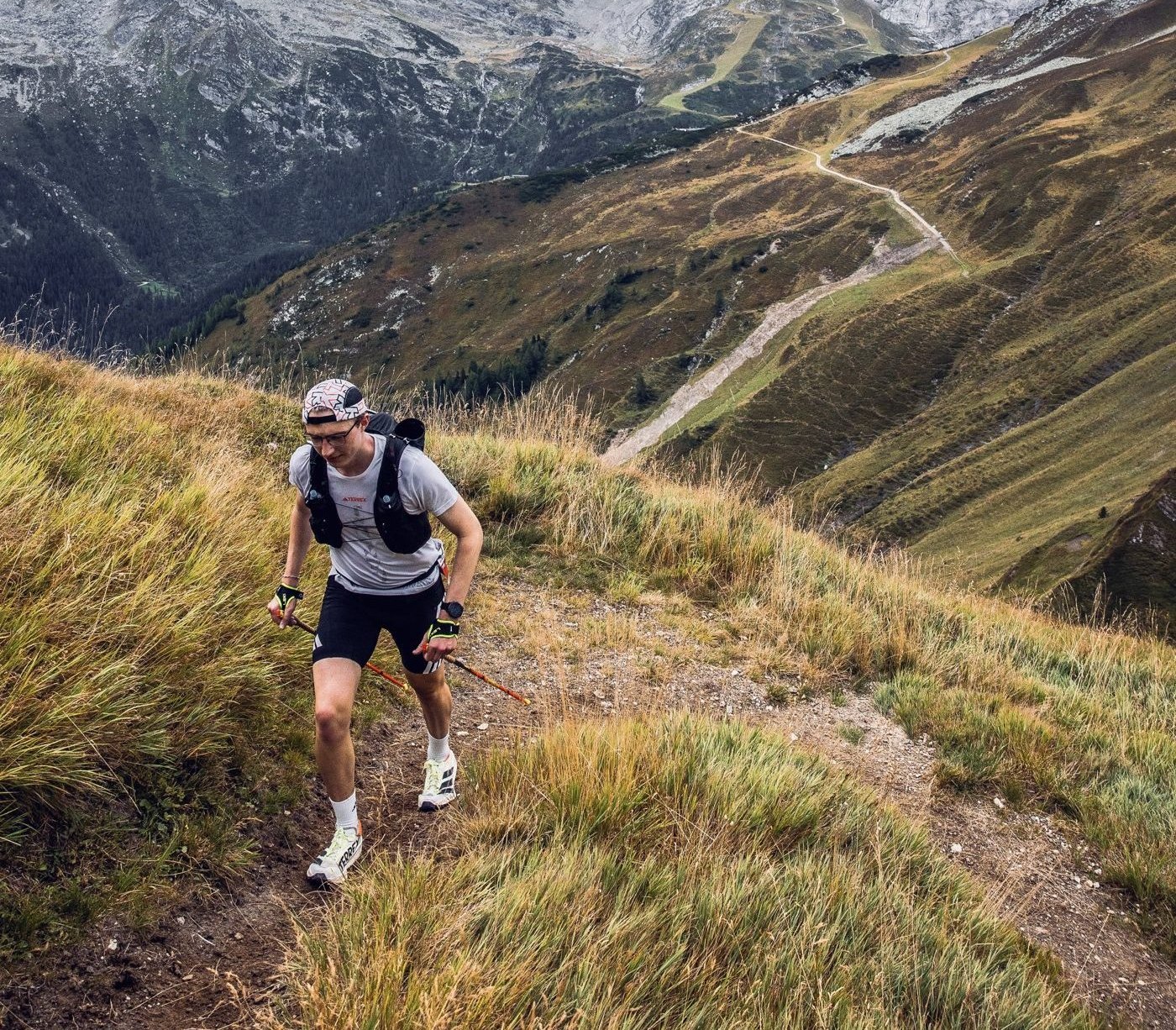 Marius läuft mit Stöcken einen Grastrail hinauf, dahinter ein Bergtal